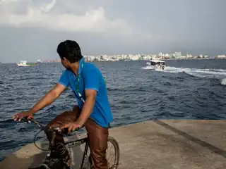 A foreign worker in Villingili gazes toward Malé while seated on a bicycle.