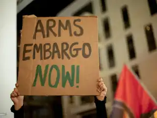 އިޒްރޭލުގެ ތަލް އަވީވްގައި ހުންނަ އެމެރިކާ އެމްބަސީ ކުރިމަތީގައި 2024 ނޮވެމްބަރު 20 ވީ ބުދަ ދުވަހު ކުރިއަށްދިޔަ މުޒާހަރާގެ ބައިވެރިއަކު، "މިހާރުން މިހާރަށް ހަތިޔާރު ފޯރުކޮށްދިނުން ހުއްޓާލާ!" ލިޔެފައިވާ ކާޑުބޯޑު ބޯޑެއް ހިފަހައްޓައިގެން.