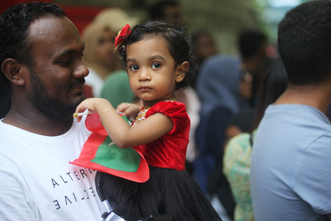 ރާއްޖޭގެ 57 ވަނަ މިނިވަން ދުވަހުގެ ޕެރޭޑް ބަލާލަން ނުކުމު ހުރި ކުޑަކުއްޖެއް ދިވެހި ދިދަ ހިފައިގެން އޭނާގެ ބައްޕަގެ އުރުގައި - ފޮޓޯ: އަހުމަދު ސިރާޖް